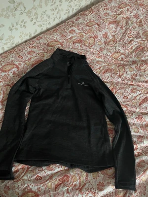 Svart långärmad träningströja Ronhill - Svart långärmad träningströja från Ronhill med halv dragkedja framtill och diskret logga på bröstet. Tillverkad i ett lätt och stretchigt funktionsmaterial som passar perfekt för löpning eller annan träning. 