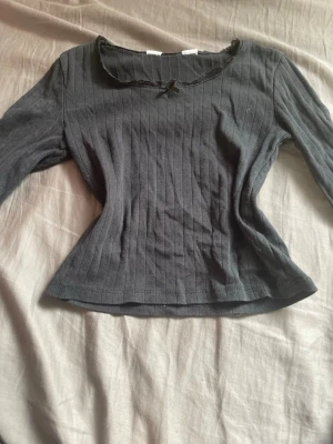 Svart ribbad blus från H&M - Snygg svart ribbad blus från H&M med lång ärm. Blusen har en liten rosett och spetsdetaljer vid halsringningen som ger en söt touch. Tillverkad i mjukt och stretchigt material som sitter bekvämt. Perfekt för dig som gillar en enkel men ändå stilren look.