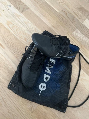 Nike Tiempo Legend 10 Elite Ag - Säljer ett par svarta Nike Tiempo legend 10 Ag (konstgräs)fotbollsskor med vita och blå detaljer. Skorna har snörning och är tillverkade i syntetmaterial med struktur för bättre bollkontroll. Perfekta för spel på gräsplan. Medföljer en svart Nike-påse. Tveka inte vid frågor🤝