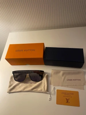 Louis Vuitton svarta solglasögon - Snygga svarta solglasögon från Louis Vuitton med rektangulär form och LV-monogram på glasen. Bågarna är i svart syntetmaterial och glasen har en mörk ton. Kommer med lfodral, putsduk och certifikat. 
