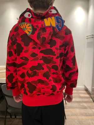Röd camo hoodie från BAPE - Säljer en röd camo hoodie från BAPE med ikoniskt hajmotiv på huvan och broderade detaljer som WGM och tiger. Hoodien har dragkedja framtill, ribbade muddar och är tillverkad i mjuk bomullsblandning. Perfekt för dig som gillar streetwear och vill sticka ut.