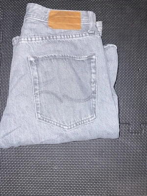 Jack & Jones jeans  - Säljer ett par ljusgrå jeans med raka ben och klassisk femficksmodell. Jeansen har en snygg läderpatch bak i midjan och är tillverkade i denim. Perfekta för en avslappnad och trendig look. W27 L30