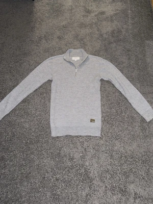 Ljusgrå stickad tröja Lindbergh - Ljusgrå stickad tröja från Lindbergh med halv dragkedja och ståkrage. Tröjan har ribbad struktur och en liten patch vid nederkanten. Perfekt för dig som gillar stilren och enkel design. Tröjan sitter åt det slimmare hållet. 