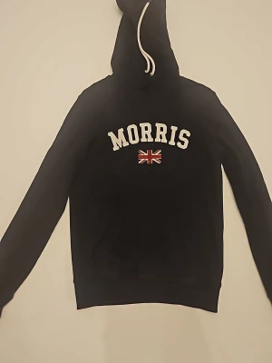 Marinblå hoodie från Morris - Snygg marinblå hoodie från Morris med vit dragsko i huvan och stor logga samt brittisk flagga på bröstet. Klassisk känguruficka framtill och ribbade muddar. Perfekt för dig som gillar stilrena och sportiga plagg med brittisk touch.