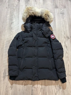 Canada Goose Wyndham  - Säljer en äkta svart dunjacka från Canada Goose med avtagbar huva och stor pälskant. Jackan har två stora fickor framtill, ribbade muddar vid ärmsluten och den klassiska Canada Goose-loggan på ärmen. Perfekt för kalla vinterdagar.