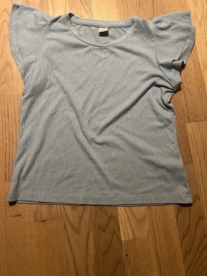 Ljusblå topp från Lindex - Snygg ljusblå topp från Lindex med korta, lite utställda ärmar och rund halsringning. Tillverkad i mjukt, stretchigt material som känns skönt mot huden. Perfekt för en avslappnad och trendig look.