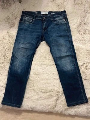 Mörkblå Replay jeans med slitningar - Säljer ett par mörkblå jeans från Replay med snygga slitningar och tvättade detaljer. Klassisk femficksmodell med dragkedsgylf. Märkespatch bak i midjan och klassiska replay loggan på fickorna.                                                                            W: 34, L: 30