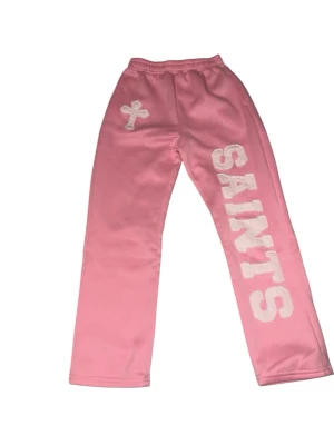 Rosa mjukisbyxor med SAINTS-tryck - Säljer ett par rosa mjukisbyxor med vitt SAINTS-tryck längs ena benet och ett kors på höften. Byxorna har resår i midjan och raka ben. Perfekta för en avslappnad och trendig look.