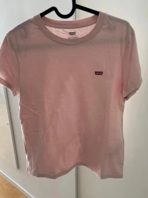 Ljusrosa Levi's t-shirt i bomull - Säljer en ljusrosa t-shirt från Levi's med klassisk rund hals och liten logotyp på bröstet. T-shirten är kortärmad och gjord i mjuk bomull, perfekt för en avslappnad och stilren look.