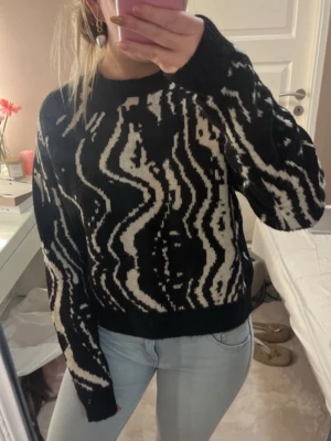 Svartvit stickad tröja med mönster - Cool svart stickad tröja med vitt abstrakt mönster. Tröjan har rund halsringning, långa ärmar och ribbade muddar. Perfekt för dig som gillar en trendig och unik look. Materialet känns mjukt och bekvämt.