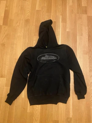 Corteiz Svart Hoodie - Först till kvarn! Svart hoodie med stor huva och magficka. På bröstet finns ett tryck med motiv av en färja eller båt i grått. Hoodien har ribbade muddar vid ärmslut och nederkant. Perfekt för en avslappnad och street-inspirerad stil.
