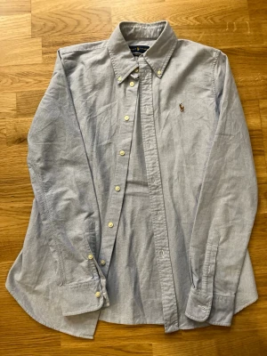 Ljusblå skjorta från Ralph Lauren - Klassisk ljusblå skjorta från Ralph Lauren med button-down krage och broderad logga på bröstet. Skjortan har långa ärmar, vita knappar och är tillverkad i mjuk bomull. Perfekt till jeans eller chinos för en clean och stilren look.