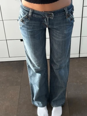 Blå bootcut jeans från D&G - Säljer ett par blå bootcut jeans från Dolce & Gabbana med låg midja och snygga detaljer som broderad bakficka och D&G-lapp. Jeansen har slitningar för en cool vintage-look och är tillverkade i klassisk denim. Perfekta för dig som gillar streetstyle.