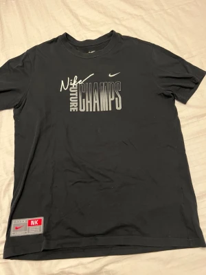 Svart Nike Future Champs t-shirt - Snygg svart t-shirt från Nike med trycket 'Future Champs' i vitt och grått på bröstet samt Nike-logga. Klassisk rund hals och korta ärmar. Perfekt för dig som gillar sportig och avslappnad stil.