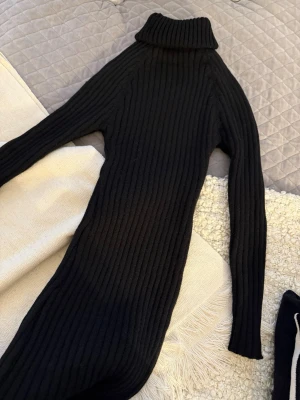 Svart ribbad stickad poloklänning - Snygg svart stickad klänning med polokrage och lång ärm. Klänningen är ribbad och har en kroppsnära passform som ger en stilren siluett. Perfekt för dig som gillar en enkel men trendig look.