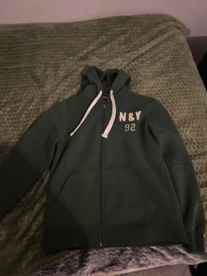 Mörkgrön hoodie från New Men - Snygg mörkgrön hoodie från New Men med dragkedja, vita snören i huvan och tryck 'N&Y 92' på bröstet. Stora fickor framtill och ribbade muddar. Perfekt för chill dagar och streetwear vibes.