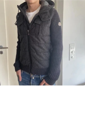 Söker - Snygg mörkgrå cardiganjacka från Moncler med stickade ärmar och vadderad framsida. Jackan har huva, dragkedja och bröstfickor med lock. Perfekt för lager-på-lager och streetstyle.