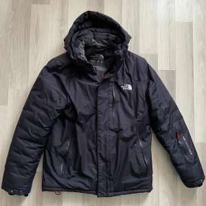 Svart dunjacka från The North Face - Säljer en svart dunjacka från The North Face med huva och dragkedja. Jackan har två sidofickor med dragkedja och en extra ficka på ärmen med The North Face-logga. Perfekt för kalla vinterdagar och riktigt skön passform.