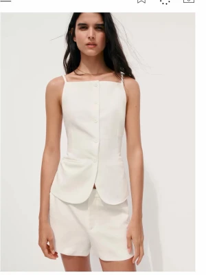 Vit linnetopp med knappar - Snygg vit linnetopp med smala axelband och knäppning framtill. Toppen har en figurnära passform och lätt utsvängd nederkant, perfekt för en stilren och fräsch look. Materialet känns svalt och luftigt.