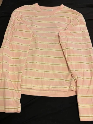 Randig långärmad topp från Monki - Söt långärmad topp från Monki i ljusrosa med tunna vita och gula ränder. Tillverkad i mjuk bomull och har en avslappnad passform. Perfekt för dig som gillar pastelliga färger och enkel, stilren design.