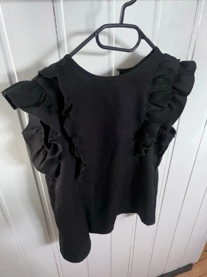 Svart ärmlös topp med volangdetaljer - Snygg svart ärmlös topp med volangdetaljer längs axlarna. Toppen har rund halsringning och en rak passform som ger en stilren look. Perfekt att matcha med jeans eller kjol för en trendig outfit. Säljer då den blivit för liten 
