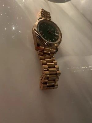 ...Guldig klocka med grön urtavla - Snygg klocka med guldigt metallarmband och grön urtavla. Klockan har romerska siffror och en markerad boettkant som ger en lyxig känsla. Perfekt accessoar för dig som vill sticka ut med stilrena detaljer.