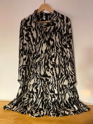 JDY XS/S mönstrad långklänning - Säljer en svartvit/beigefärgad långklänning med abstrakt zebramönster. Klänningen har lång ärm, krage och en lös passform med volangdetaljer. Tillverkad i ett mjukt och lätt tyg, perfekt för dig som gillar statementplagg. Slutar strax under knäna. Året-om-klänning som passar lika bra till bara ben som strumpor/strumpbyxor. Elegant känsla. Från JDY i storlek 34 (oversized modell). Använd en gång!