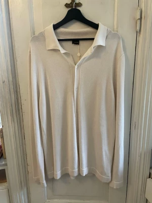 Vit cardigan från Rose & Born - Klassisk vit cardigan från Rose & Born med krage och knäppning framtill. Tillverkad i ett mjukt, tunt stickat material som ger en clean och stilren look. Perfekt att bära över en t-shirt eller skjorta för en avslappnad men snygg stil.