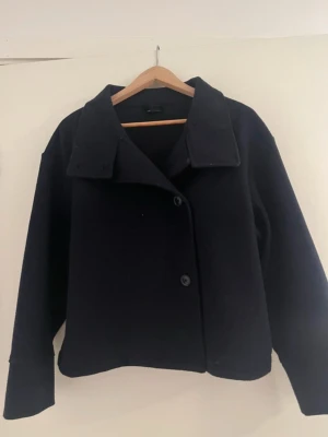Svart marinblå kort kappa -  svart marinblå kort kappa i ull med bred krage och knäppning framtill. Modellen är kortare och har raka ärmar samt minimalistisk design utan synliga fickor. 
