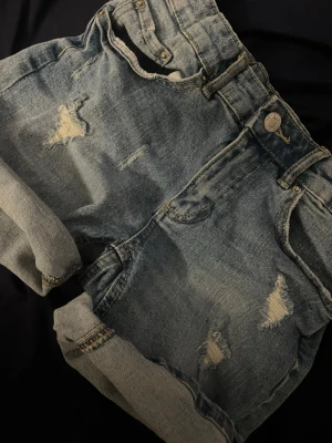 Lågmidjade jeansshorts med slitningar  - Säljer ett par blå denimshorts med uppvikta benslut och snygga slitningar framtill. Klassisk femficksmodell med knapp och dragkedja i midjan. Perfekta för sommaren