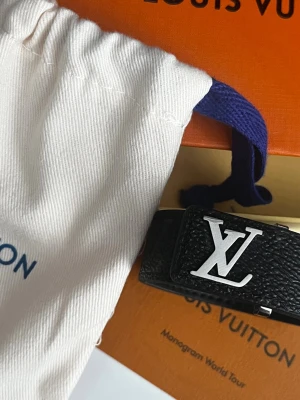 Louis Vuitton svart läderarmband - Säljer ett svart armband från Louis Vuitton i exklusivt läder med silverfärgad LV-logga framtill. Armbandet har en stilren och modern design med diskret monogrammönster i lädret. Perfekt för dig som vill ha en lyxig accessoar med ikonisk branding.