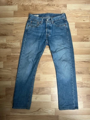 Levi's blå 501 - Klassiska blå jeans från Levi's med rak passform och fem fickor. Jeansen har en ljus tvätt och detaljer som kopparfärgade nitar och läderpatch bak i midjan. Tillverkade i slitstark denim, perfekta för en avslappnad och tidlös stil. Mått finns hör av dig vid frågor:)