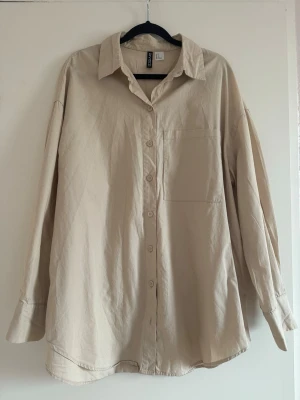 Beige oversized skjorta från H&M - Stilren beige skjorta från H&M i oversized modell. Skjortan har klassisk krage, knäppning framtill och en bröstficka. Tillverkad i mjuk bomull och har långa ärmar med manschett. Perfekt för en avslappnad och trendig look.