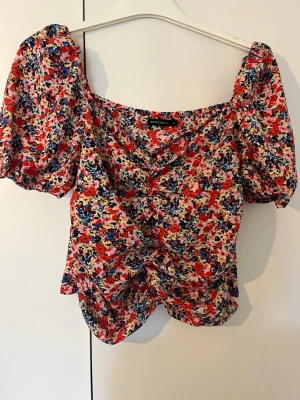 Blommig topp med puffärm från Rut & Circel - Superfin blommig topp från Rut & Circel med korta puffärmar och v-ringning. Toppens tyg är lätt och har ett färgglatt blommönster i rött, blått och vitt. Snygg rynkdetalj framtill och croppad passform. Perfekt för dig som gillar färg och trendiga detaljer.