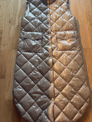 Glansig silverfärgad dunväst - Snygg lång dunväst i glansigt silver med quiltat rutmönster och stora fickor framtill. Västen har hög krage och stängs med guldfärgade tryckknappar. Perfekt för lager på lager och en cool streetstyle-look.