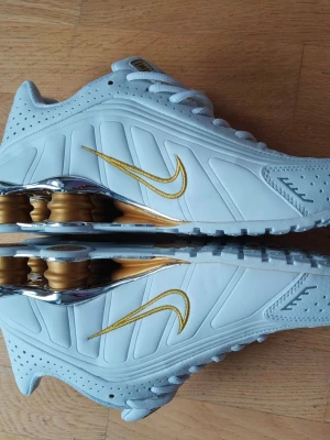 Nike Shox vita och guld sneakers - Nike Shox sneakers i vitt med guldfärgade detaljer och metallic guld Shox-dämpning i hälen. Skorna har en låg silhuett, perforerade partier och ett guldigt Swoosh på sidan. Tillverkade i syntetmaterial och mesh för en sportig look. 