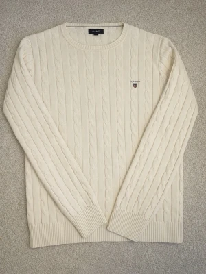 Vit kabelstickad tröja från Gant - Kabelstickad tröja i mjuk vit bomull från Gant. Klassisk rund halsringning, ribbade muddar och diskret broderad logotyp på bröstet. Perfekt för lager-på-lager och en stilren look.