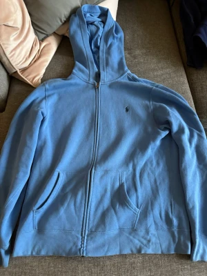 Blå hoodie från Polo Ralph Lauren - Snygg blå hoodie från Polo Ralph Lauren med dragkedja framtill och två fickor. Klassisk huva och den ikoniska lilla loggan broderad på bröstet. Perfekt för chill dagar eller när du vill ha en enkel men stilren look.