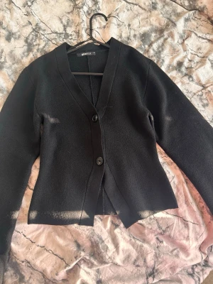 Knitted sculptured cardigan  - Stilren svart kofta från Gina Tricot med två stora knappar framtill och v-ringad hals. Koftan har lång ärm och tajt passform kring midjan. Materialet känns mjukt och lätt, vilket gör den enkel att bära till olika outfits.