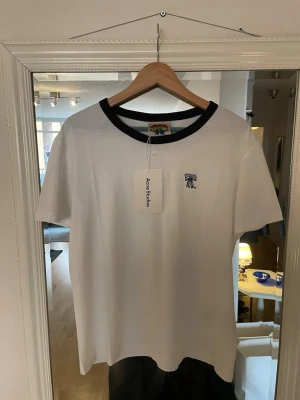 Vit t-shirt  Acne Studios - Vit t-shirt från Acne Studios med svart kontrastkrage och liten broderad logga på bröstet. Klassisk passform och korta ärmar. Tillverkad i mjuk bomull som känns skön mot huden. Perfekt för en clean lite cropped och stilren look. Storlek M. Ny pris 1300kr