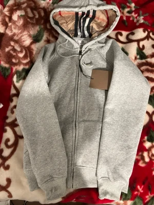 Grå zip-hoodie från Burberry - Snygg grå hoodie från Burberry med dragkedja och klassiskt rutigt foder i huvan. Tröjan har ribbade muddar, dragsko i huvan och två fickor framtill. Perfekt för en stilren och avslappnad look.