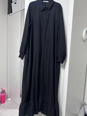 Svart långklänning med volangkant - Stilren svart långklänning med klassisk krage, knäppning hela vägen framtill och långa ärmar med lätt puff vid mudden. Klänningen har en volangkant nedtill som ger en trendig touch. Perfekt för dig som gillar enkel men snygg stil.