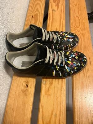 Maison margelas  - Unika svarta sneakers från Replica med färgstänk i gult, blått, rött och vitt över hela ovandelen. Skorna har beige snörning och innersula, samt klassisk låg siluett. Tillverkade i läder med gummisula. Perfekta för dig som vill sticka ut med din stil. Priset är ej hugget i sten !!!