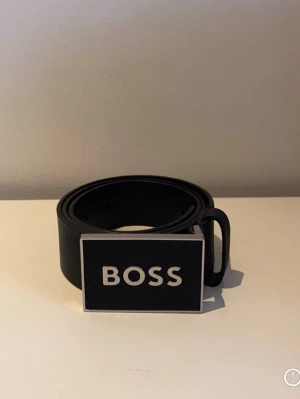 Svart BOSS bälte med stor logga - Snyggt svart bälte från BOSS med en stor, rektangulär metallspänne där BOSS-loggan syns tydligt. Bältet har en stilren och modern look, perfekt för att lyfta vilken outfit som helst.