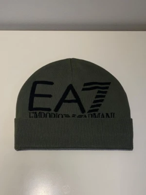 Olivgrön EA7 mössa från Emporio Armani - Snygg olivgrön mössa från Emporio Armani EA7 med svart logotyp framtill. Tillverkad i 100% akryl med ribbstickad kant. Perfekt accessoar för att lyfta din streetwear-look.
