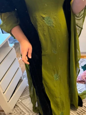 Grön kurdisk långklänning i siden - Säljer en elegant grön långklänning i siden med diskreta broderade detaljer och löst fall. Klänningen har långa ärmar och en klassisk, rak siluett som ger en tidlös look. Perfekt för dig som vill sticka ut med en unik färg och exklusivt material.