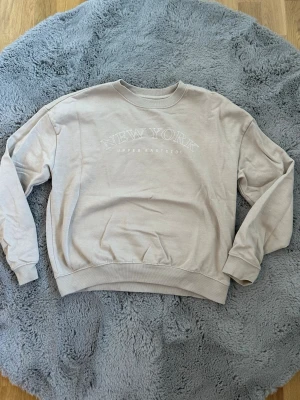 Beige sweatshirt med New York-tryck - Beige oversized sweatshirt med texten 'NEW YORK UPPER EAST SIDE' tryckt framtill. Tröjan har rund halsringning, långa ärmar och ribbade muddar vid ärmslut och nederkant. Perfekt för en avslappnad och trendig stil.