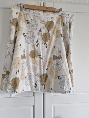 Esprit Vit blommig kjol i bomull - Säljer en vit kjol med beige och brunt blommönster. Kjolen har en lätt utställd form och är gjord i mjuk bomull, perfekt för varma dagar. Blommorna är stora och detaljerade vilket ger kjolen en fräsch och somrig känsla. Spetskant i nederkant och band o sidan. Dragkedja i sidan
