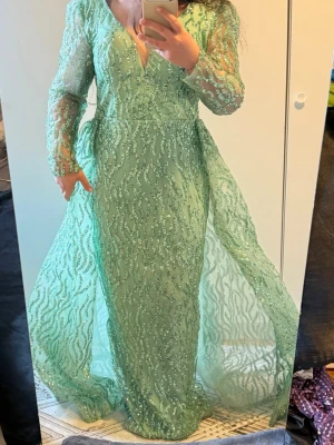 Mintgrön långklänning med glitterdetaljer - Superfin mintgrön långklänning med transparenta ärmar och glittriga mönster över hela klänningen. Djup v-ringning och lätt puffiga axlar ger en elegant look. Perfekt för bal eller festliga tillfällen.