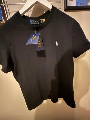 Svart t-shirt från Polo Ralph Lauren - ( Pris kan diskuteras)Klassisk svart t-shirt från Polo Ralph Lauren med den ikoniska broderade loggan på bröstet. T-shirten har rund hals och korta ärmar, tillverkad i mjuk bomull för en skön och stilren look. 